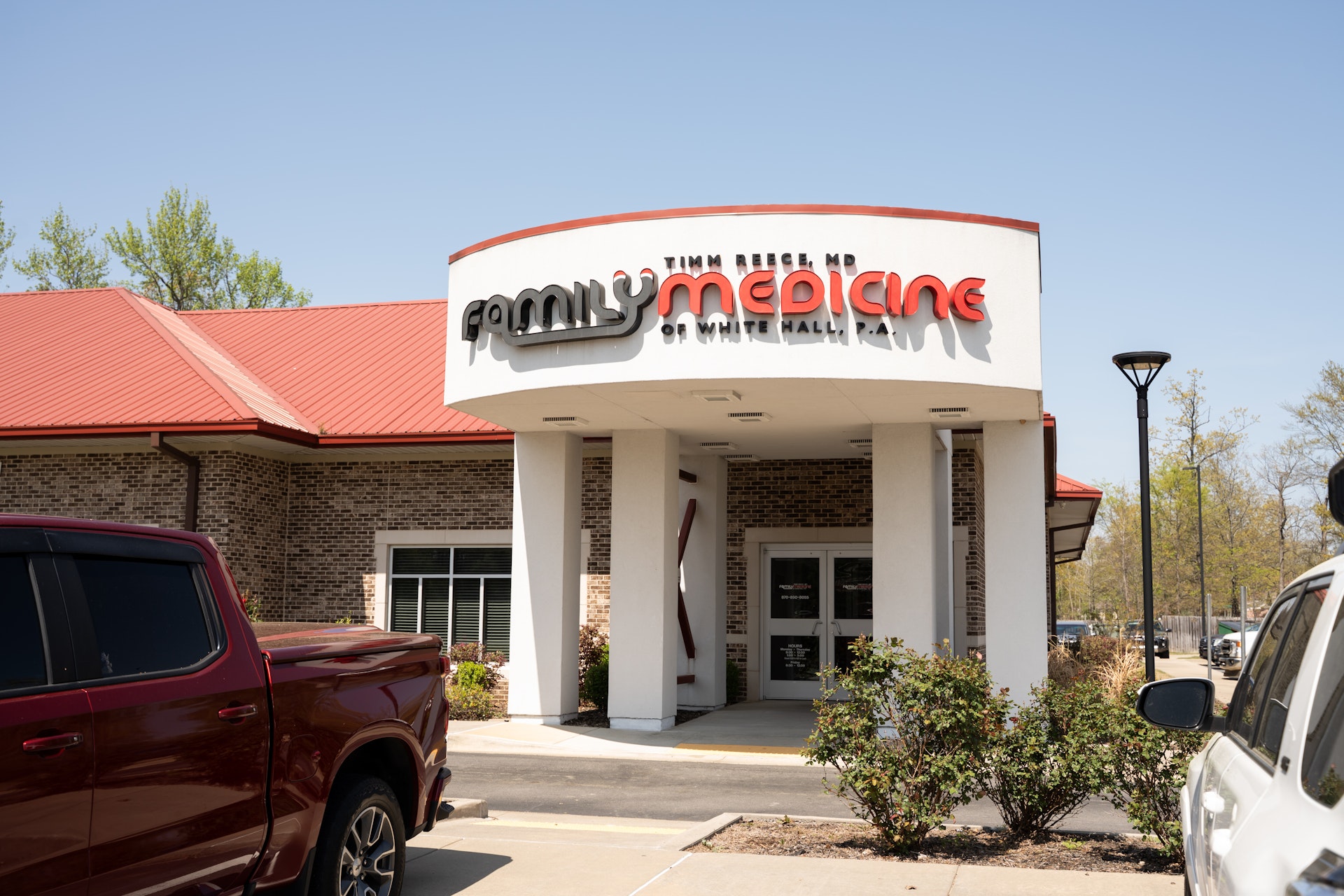 A medical clinic building with a sign reading Family Medicine, Tim Meese MD, 60 White Hall Rd. The clinic has a red roof, white columns, and is surrounded by parked vehicles and greenery.