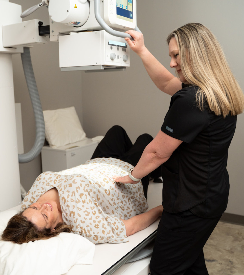 A healthcare worker positions a woman for an X-ray exam; the woman lies on her back on the examination table, wearing a hospital gown, while the healthcare worker adjusts her position and operates the X-ray machine.