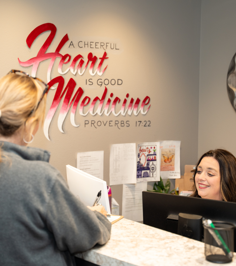 Family Med of WH-085 resized A woman at a reception desk speaks with a receptionist who is smiling. Behind them, a wall sign reads, A cheerful heart is good medicine - Proverbs 17:22. Papers and notes are pinned to the wall.