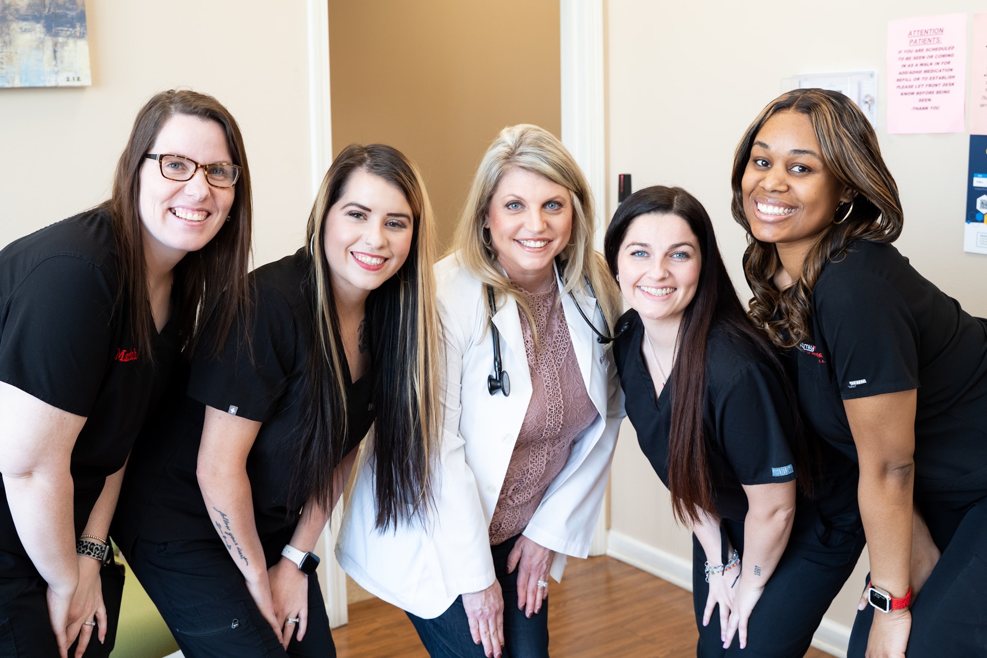Family Med of WH-118 Five smiling women, four in black scrubs and one in a white coat with a stethoscope, pose together indoors in a medical or clinical setting. They stand closely and lean forward, looking at the camera.