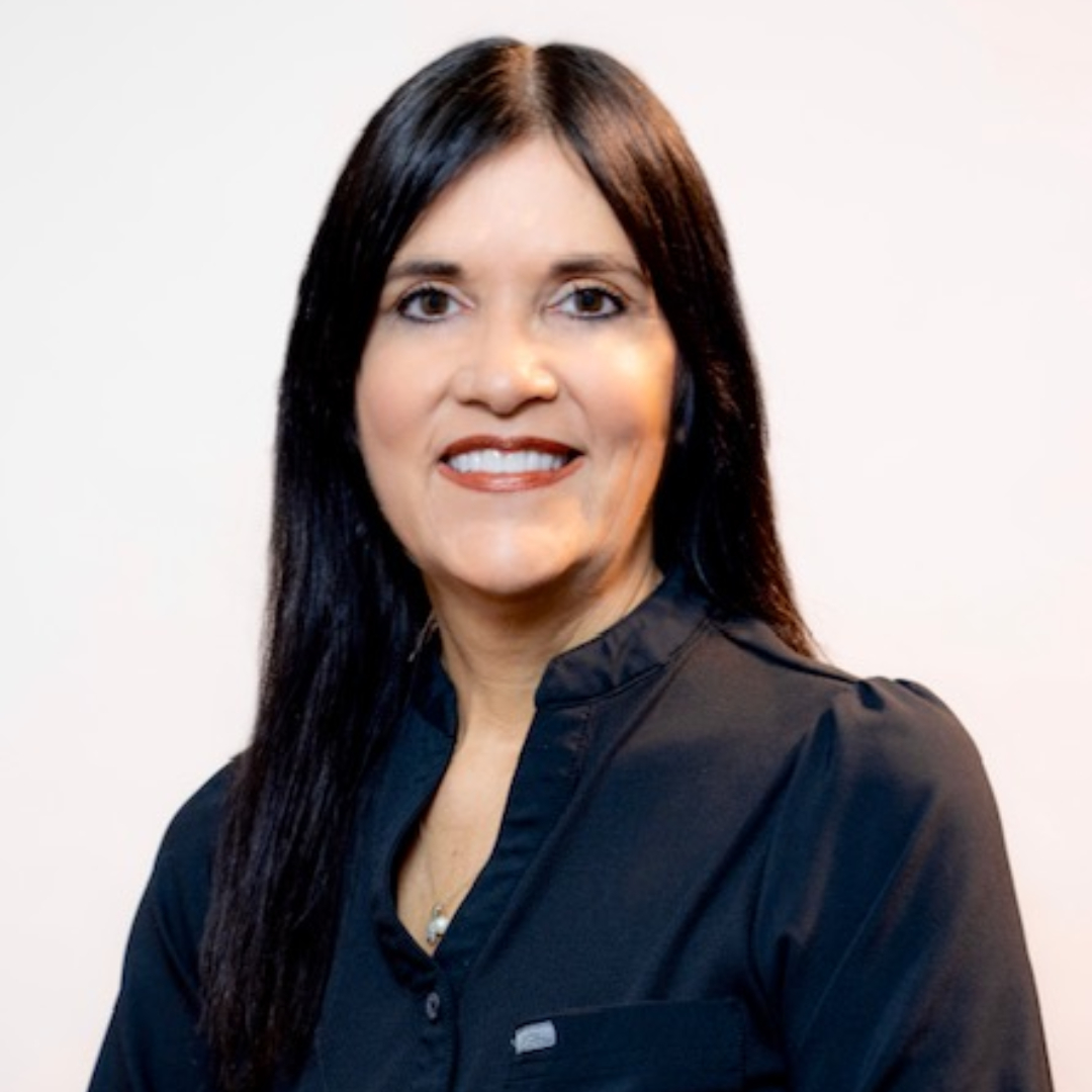 Johnnie Kelsey with long straight dark hair, wearing a dark blouse, smiles at the camera against a plain light background.
