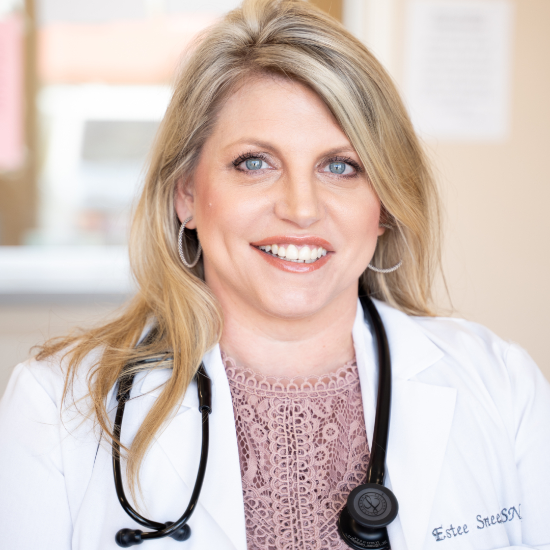 Estee Sneed, APN with long blonde hair, wearing a white medical coat, pink lace blouse, and a stethoscope around her neck, sits indoors with a blurred background.