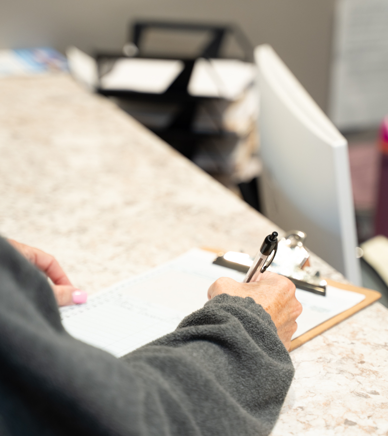 A person wearing a gray sweater is writing on a clipboard with a pen while standing at a marble countertop. The background is blurred, showing an office or reception area.