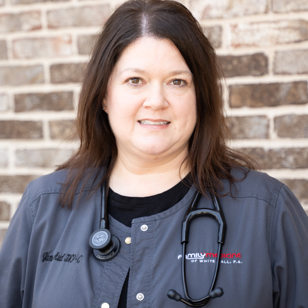 Jamie Aud, APN with long brown hair wearing a gray medical uniform and a stethoscope around her neck stands in front of a brick wall, smiling slightly at the camera.