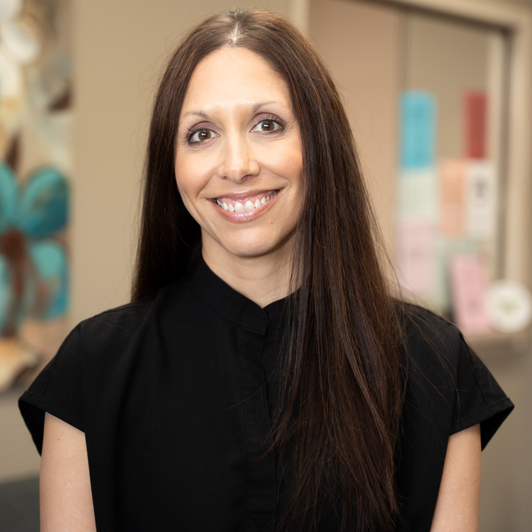 Karla Young with long straight brown hair, wearing a black top, smiles at the camera. She stands indoors, with a blurred background featuring colorful posters and artwork.