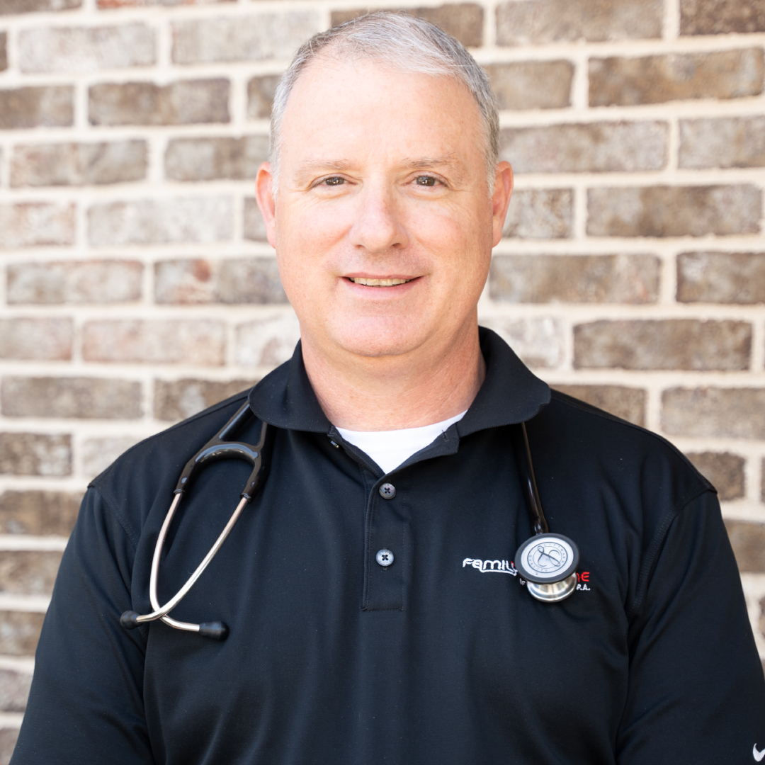 Timmothy C. Reece, MD with short gray hair wears a black polo shirt and has a stethoscope around his neck, standing in front of a brick wall.